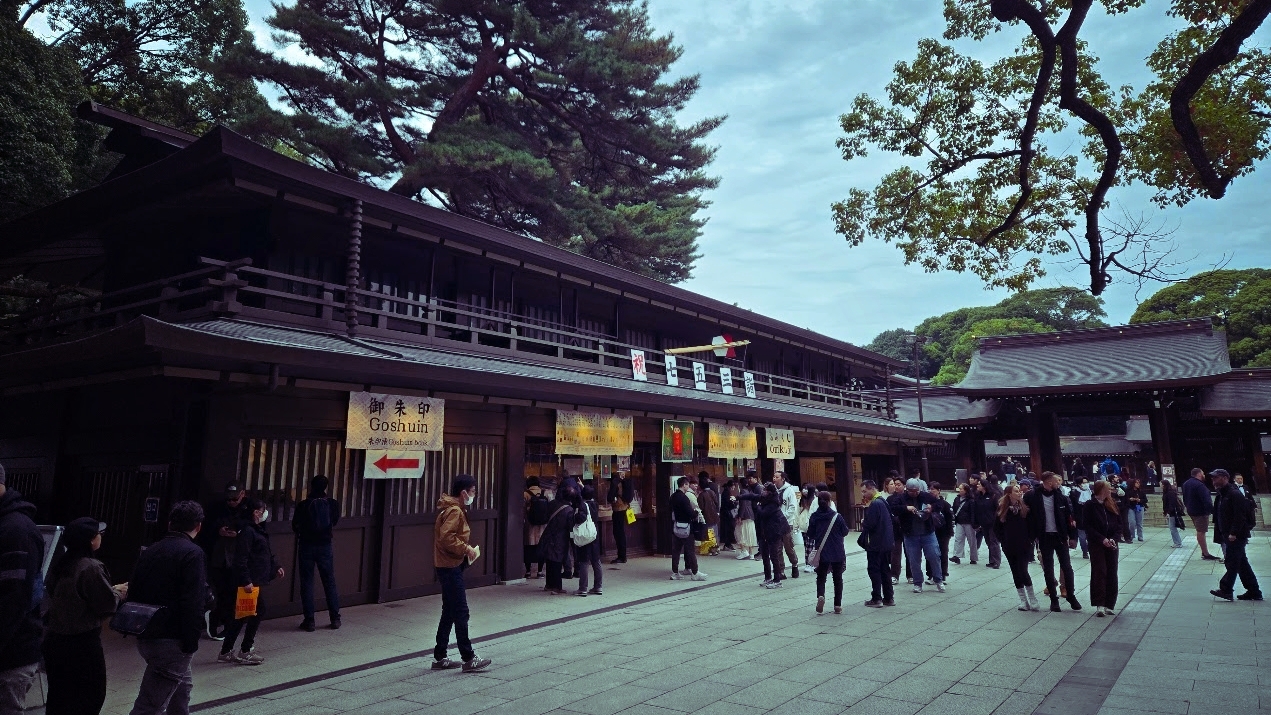 A photo of a crowd in a courtyard near another temple building, with a small sign pointing towards the back of a building, with the sign saying the word 'goshuin' on it.