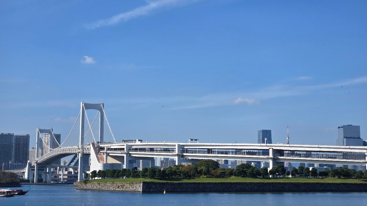 A photo of a large white bridge with one end near the camera, and the other end across the other end of a span of water to reach Tokyo's central business district.
