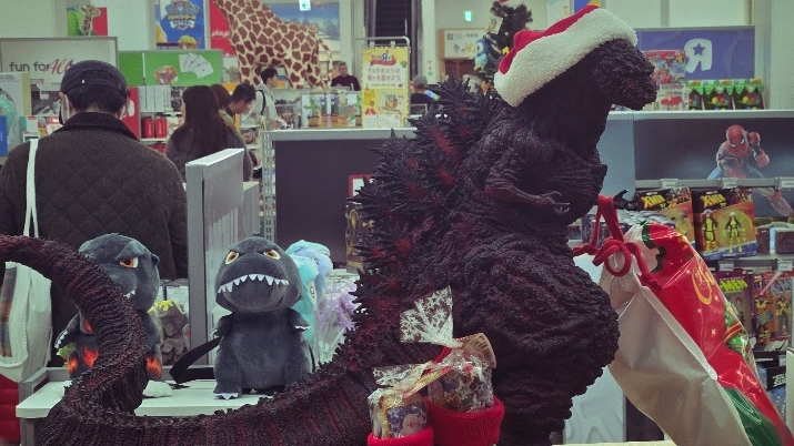 A photo of a dark blackish-red Godzilla wearing a Santa hat. Yes, this Godzilla spews blood and has flesh sizzling and melting off of it - it's not the version of Godzilla you'd expect to see promoting a family holiday.