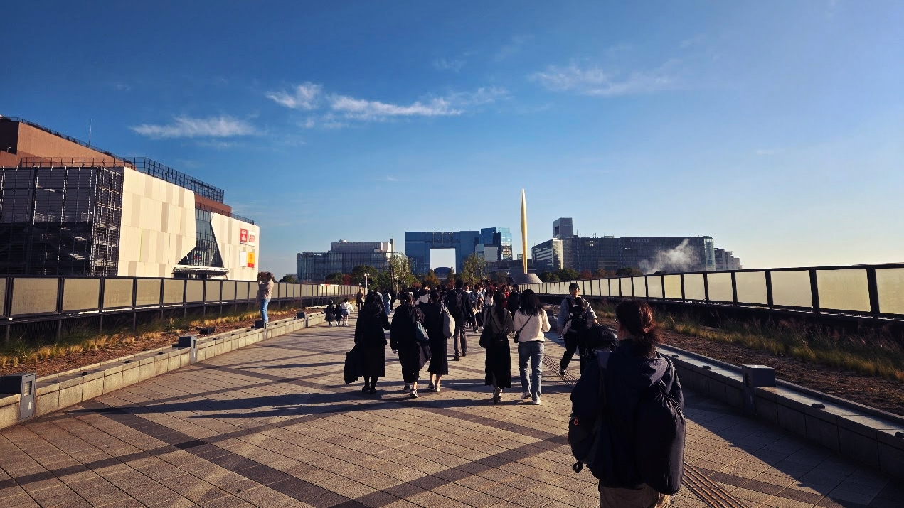 A photo of a crowd walking across a large pedestrian bridge, with wide open skies above and giant shopping complexes at the end of the bridge.