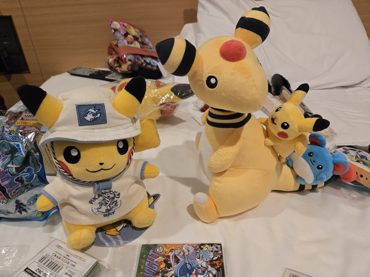 A photo of some merch from the Pokemon Center Tokyo Bay store, including two different location-exclusive plushies featuring a Pikachu wearing a bucket hat and sunshirt and an Ampharos carrying a Marill and Pikachu on its back.
